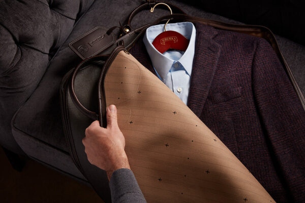 A hand opens a GHURKA® Weekender Bag, revealing the interior beige lining and repeated black aircraft motif design. Beneath the bag lies a light blue collared shirt and burgundy woven plaid jacket.